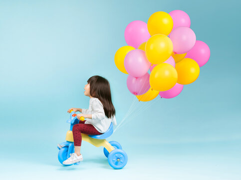 Little girl on a kids tricycle with colorful balloons behind on plain background.