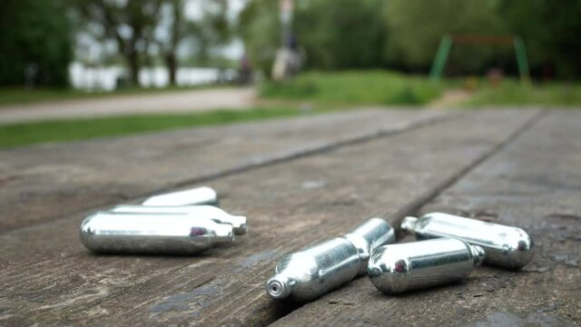 Closeup pile of chrome nitrous oxide recreational drug cylinders on wooden park table dolly left