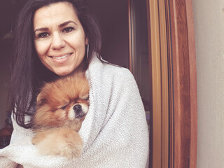 Smiling woman hugging  cute fluffy  pomeranian spitz dog wrapped in white blanket  looking  out the window on a   cold winter day.  