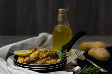Baked potato wedges with rosemary, garlic and lemon in a small pan. Raw potatoes, vintage silver fork, rosemary and olive oil in the background. Rustic and dark aesthetic 