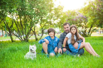 Fototapeta premium Dad with daughter and son are sitting on the grass in the park, daughter is on the mans knees. A boy in shorts and a denim jacket. In the background are trees. Nearby is a cute white dog.