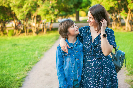 Mom With Teenage Son Are Walking In The Park. They Look At Each Other And Smile. A Woman Hugs Boy And Straightens Her Hair. The Concept Of Mother's Day, Relationships With Adolescents, Care And Love.