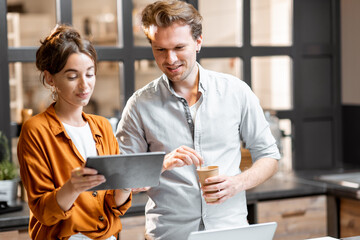 Two young managers or shop owners having some discussion while standing with a digital tablet at the counter of the shop or cafe. Small business management concept