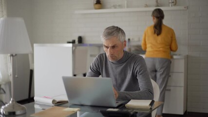 Tracking medium shot of senior man working on laptop computer and thinking sitting at table at home while his wife washing dishes in kitchen - Powered by Adobe