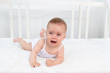 Fototapeta premium baby girl 6 months old lying on the bed in the children's room on her stomach and crying, baby morning, baby products concept