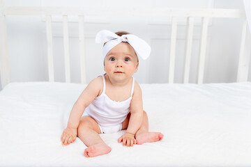 smiling baby girl 8 months old sitting in a crib in a children's room in white clothes and a bow and looking at the camera, morning baby, baby products concept