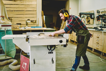 Young craftsman concentrated on carpentry work with wooden using circular for saw plank, skilled male joiner in wear for safety and protection during using professional equipment in workshop.