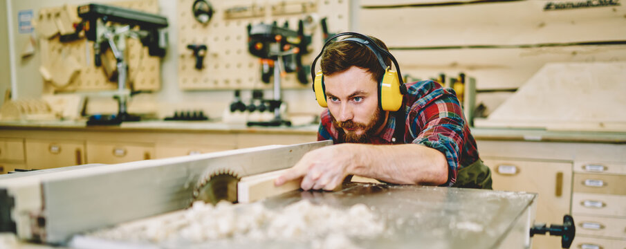 Craftsman concentrated on carpentry work with wooden using circular for decorative plank, skilled male joiner in headphones for safety and protection during using professional equipment in workshop