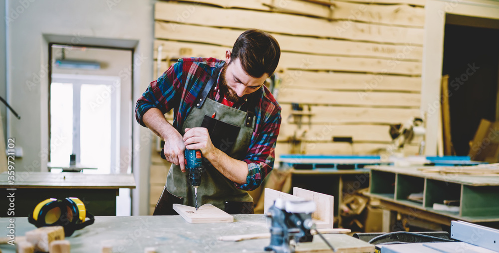 Wall mural skilled craftsman in apron using drill while working with wooden plank for furniture in own workshop