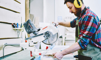Professional male carpenter using modern electronic carpenter during woodworks in workshop, skilled young guy learning to use equipment on artisan courses dressed in eyewear and headphones for safety