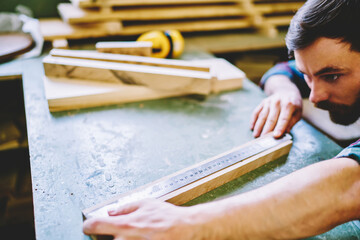 Cropped image of caucasian male student of handmade carpentry courses measure details with ruler on working place; skilled concentrated; foreman using tools for checking bricks and planks in workshop