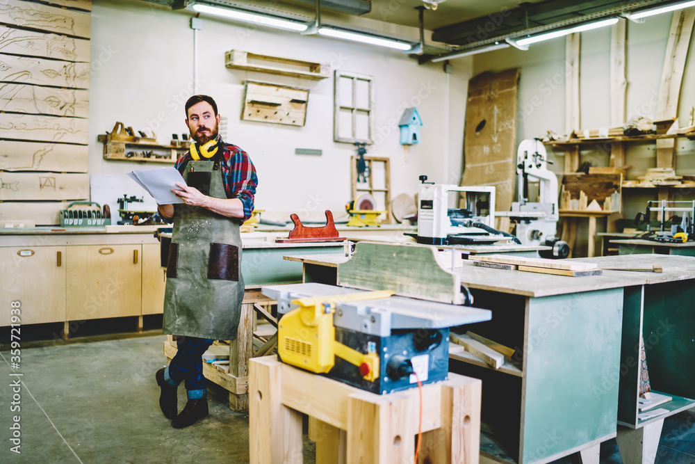 Wall mural pensive male engineer in apron holding paper for making sketch and planning working in own workshop 