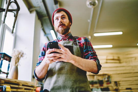 Caucasian bearded woodworker in apron holding mobile phone dialing number for making call, skilled male owner of manufacture workshop chatting with clients via smartphone during working day .