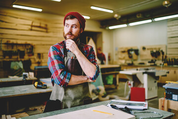 Dreamy handsome male artisan in apron pondering on ideas for making wooden stuff looking away in...
