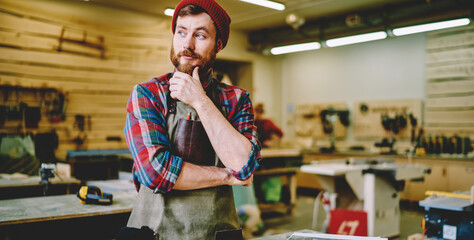 Bearded craftsman in apron pausing to think at workshop