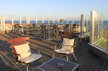 Rooftop cafe with panoramic sea views. Outdoor terrace with a glass fence around the perimeter. Modern bar furniture: dark rattan tables and chairs and stylish wooden chairs with white pillows.