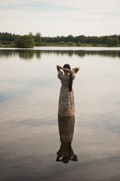 Girl in classic vintage long dress standing in water of lake seen from behind doing her long hair in evening light