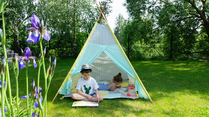 Girl draws with crayons in a tipi patchwork during the summer holidays. A schoolboy recently learned to read and carefully reads a children's book in a flower garden on a lawn outside the city © nieriss
