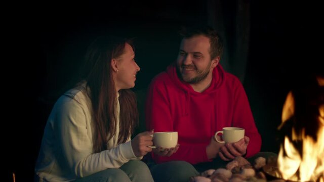 Cute millennial couple flirting while drinking hot tea by fire on patio, beautiful gilfriend and boyfriend enjoying romantic weekend near campfire at night. Nice conversation, cozy evening, dream