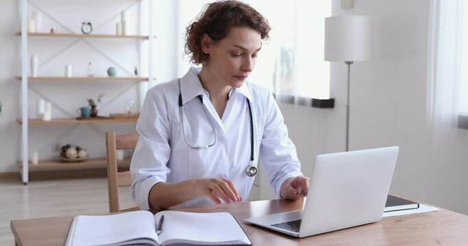 Focused 30s female general practitioner in white uniform sitting at workplace, doing paperwork and using medical software application on computer, involved in research or making report at clinic.