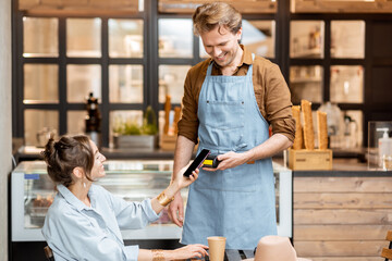 Happy waiter with a client at the cafe or ice-cream shop, making contactless payment with a smartphone