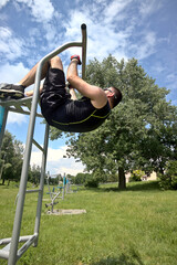 A young man in sports outfit trains abdominal muscles at an outdoor gym in the city park