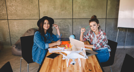 Portrait of two female students dressed in stylish cassual clothing sitting at wooden table with laptop computer in wifi zone .Smart hipster girls studying in coworking space while looking at camera