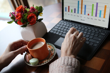 Home based remote work during new reality and quarantine, Social distancing concept.Working space view with cup of coffee with sweat treats between woman hands, working on the laptop computer and vase