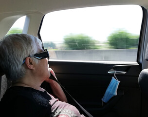 an elderly woman enjoying the scenery outside the car window with her white hair blowing in the wind with detail of anti COVID mask