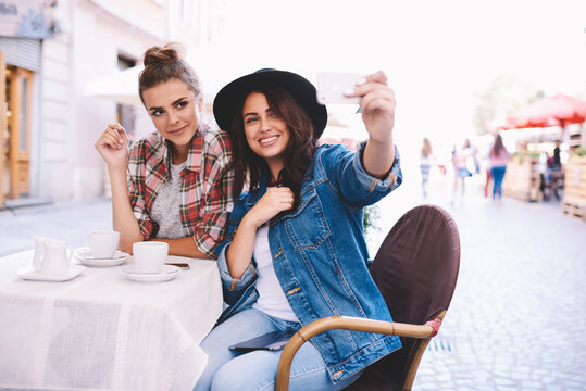 Two charming female dressed in trendy clothing making selfie and updating profile image in social network.Good looking young woman spending free time in sidewalk cafe and taking photo on smartphone