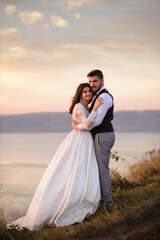 The bride and groom are hugging on the background of the lake during sunset.