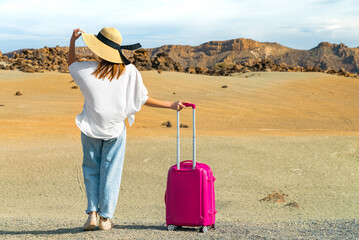 Girl stand in a white shirt and hat with a pink suitcase on a volcanic sandy scenery. Teide volcano, Tenerife, Spain.