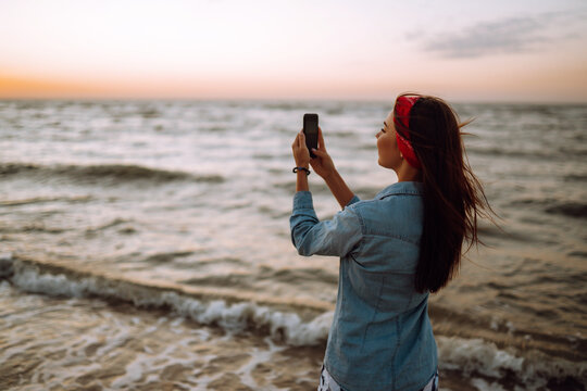 Young girl sitting on the beach photographs the sea at sunset. Woman on vacation using mobile phone to take photo of sea waves.