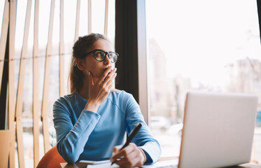 Professional pretty journalist in trendy spectacles pondering about ideas for article publication and writing main details into notepad.Pensive young hipster girl spending free time sitting in cafe