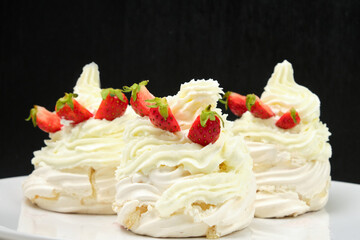 Delicious light meringue cakes with cream decorated with fresh halves of strawberries on a black background. Tasty homemade pastry. 