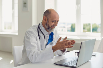 Senior doctor speaks with patient using laptop online video webinar consultation sitting in clinic...