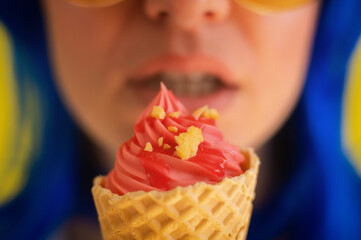 Closeup of sensual female lips with a waffle cone of pink ice cream. Cropped photo of a woman in sunglasses with blue hair happily eating pink gelato on a hot summer day. Macro. Happy sweet tooth.