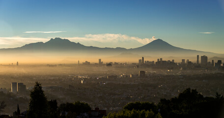 Panoramic view of Mexico City at dawn, with fumarole of active volcano Popocatepetl beside Iztaccihuatl, mist between skyscrapers