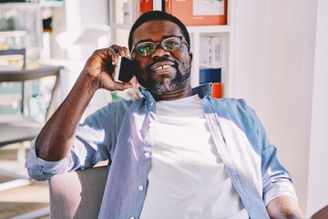 Positive young dark skinned male having cellular conversation during leisure time, handsome smiling african american hipster guy making telephone call satisfied with tariffs and mobile connection .