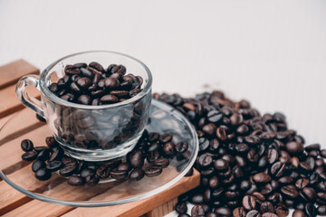 coffee cup and saucer  filled with black coffee beans, both placed on a wooden platform with a heap of coffee beans beside it. white background