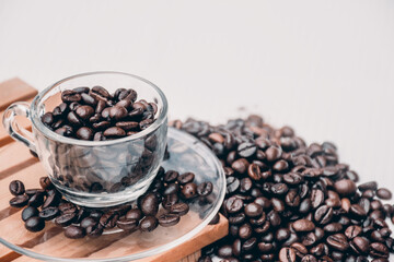 coffee cup and saucer  filled with black coffee beans, both placed on a wooden platform with a heap of coffee beans beside it. white background