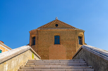 Stairs of brick stone bridge across water canal and Saint Domenico catholic church in Chioggia town historical centre, blue sky background in summer day, Veneto Region, Northern Italy