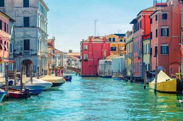 Fototapeta na wymiar Chioggia cityscape with narrow water canal Vena with moored multicolored boats between old colorful buildings and brick bridge, blue sky background in summer day, Veneto Region, Northern Italy