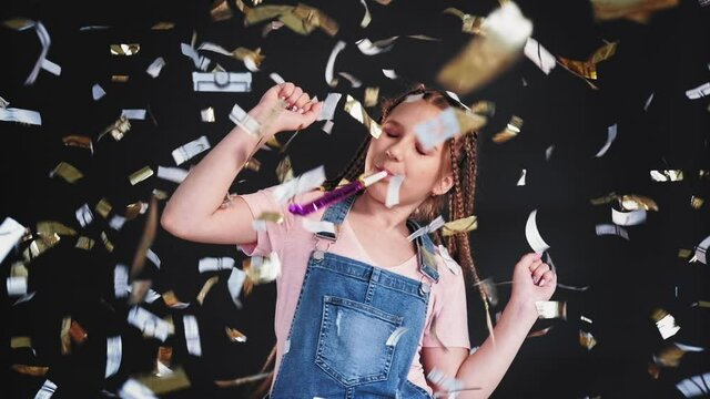 Festive portrait. Birthday party. Teen girl dancing blowing horn in confetti rain isolated on black background.