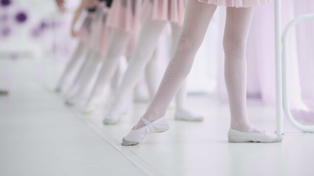 Close up feet of unrecognizable little girls in white socks and ballet shoes, their ballet teacher correcting them