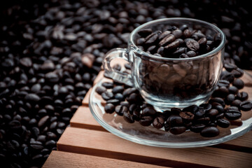 glass coffee cup and a saucer both filled with coffee beans, with black coffee beans in the background