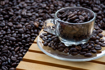 glass coffee cup and a saucer both filled with coffee beans, with black coffee beans in the background