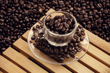glass coffee cup and a saucer both filled with coffee beans, with black coffee beans in the background