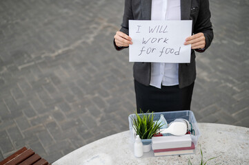A faceless business woman is fired and stands outdoors with a box of personal items holding a sign...