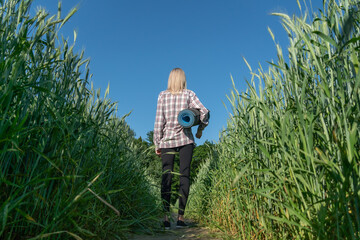young blonde woman in black leggings and a shirt walks through a field of green rye into the woods for training and holds a sports Mat in her hands
